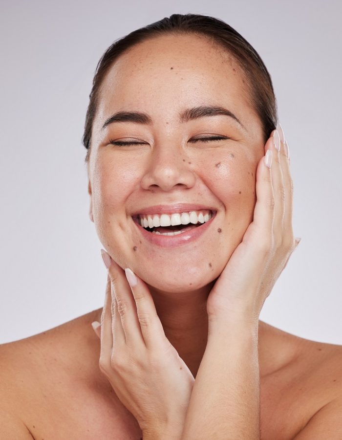 Skincare, hands on face and woman laughing in studio for cosmetics, dermatology and beauty glow. Happy aesthetic asian model person with makeup, skin mole and facial self care on grey background.